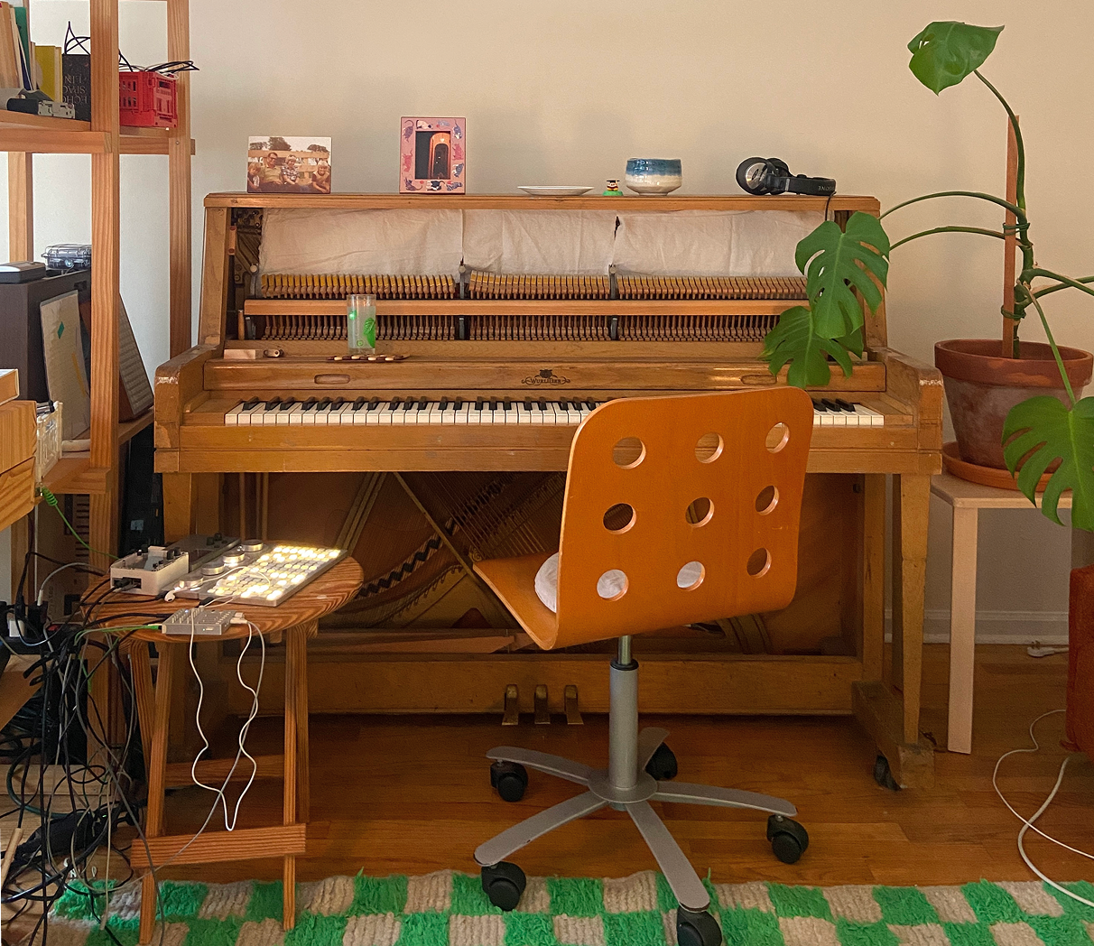 A photo of a piano in a cozy room surrounded by a plant, some electronic music equipment and wooden furniture